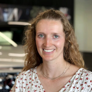 Foto der Recruiterin Judith Schrott mit freundlichem Blick vor einem verschwommenen Autohaushintergrund.